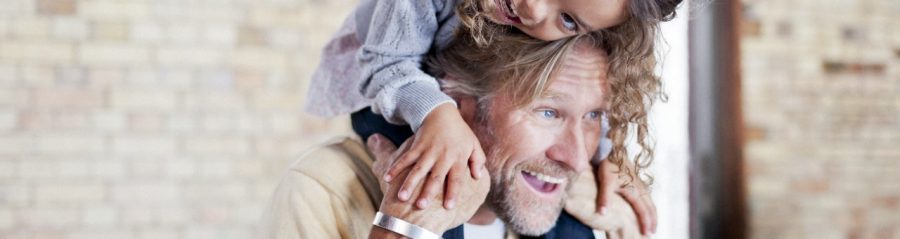 Close-up of a father and his girl fooling arrounding; her climbing his back; both hayppily smiling, unplastered brick wall in the back