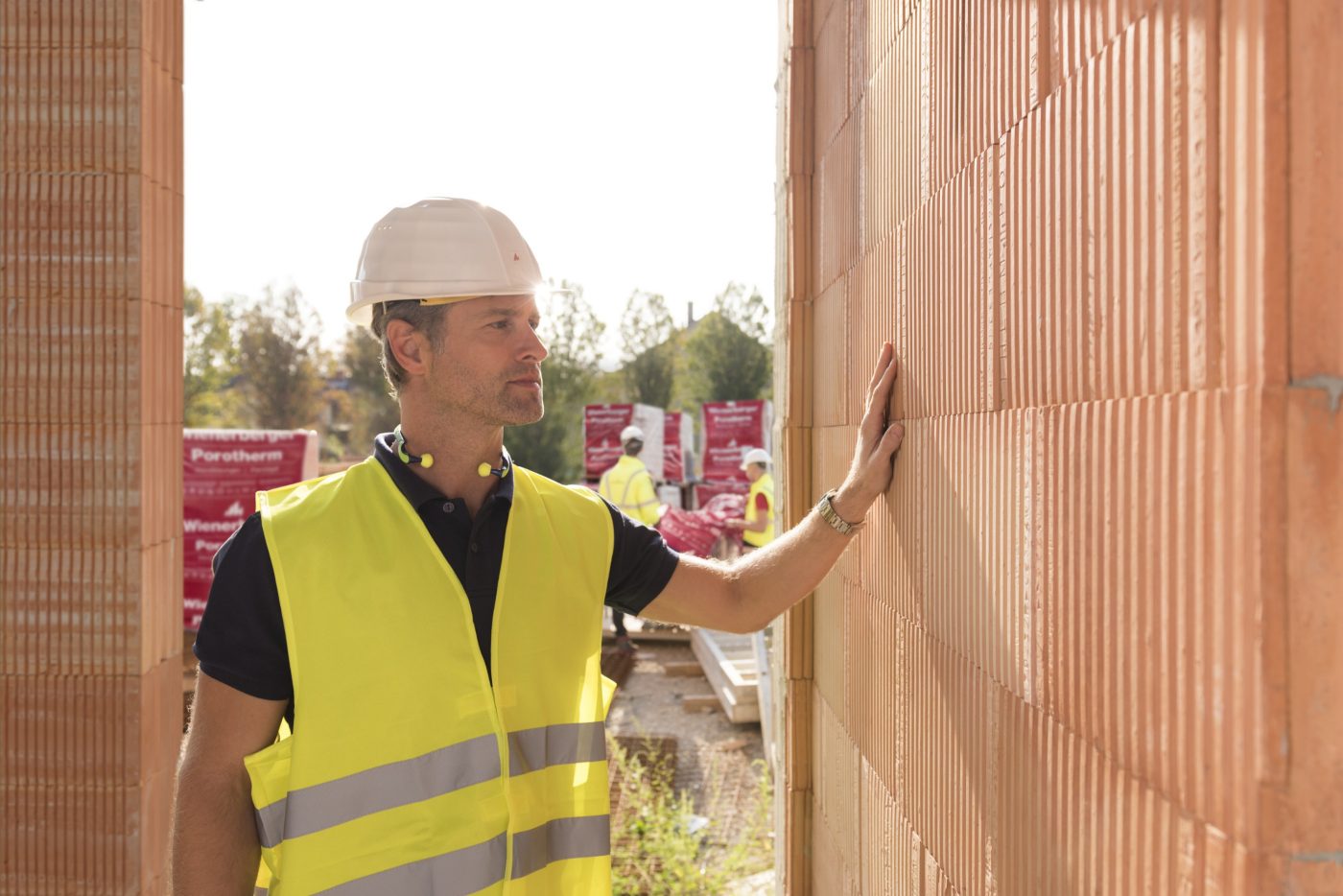 Builder on construction site touching clay block wall, construction workers and clay block pallets in the background