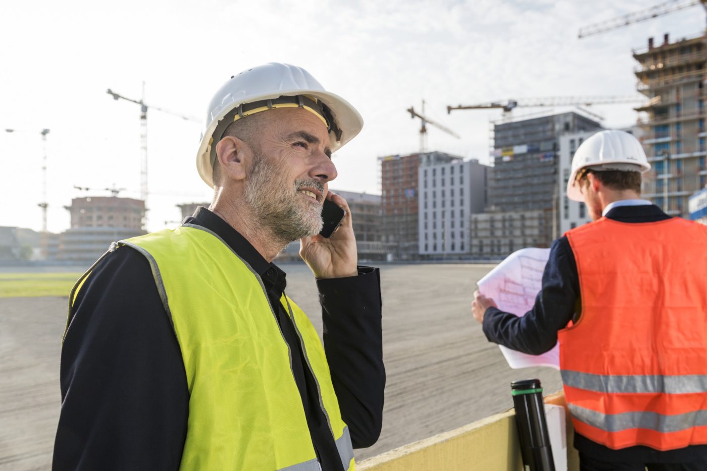 Architect on the phone while his colleague is looking at a plueprint, unfinished buildings in the background, Fast Forward Commercial Excellence